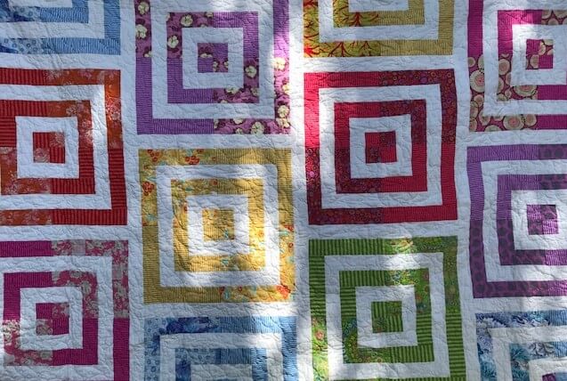 A quilt of concentric square blocks in bright prints and white, hanging outdoors under dappled tree shadows