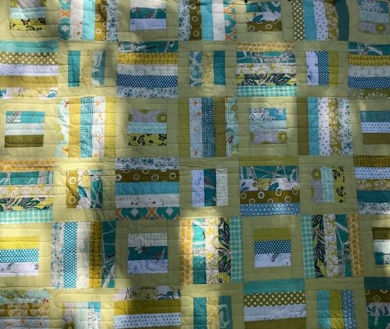 A patchwork quilt of green, teal and yellow striped blocks set in pale olive sashing